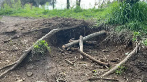 An open hole in a footpath about a foot deep, with branches and roots strewn across it.