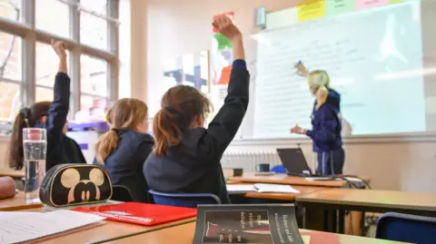 A classroom setting where a teacher is writing on a whiteboard and three pupils are sitting at their desks, two of which have their hands up.