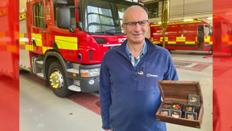 A man in a blue 1/4 zip fleece, glasses and a blue checked shirt satnds infront of fire engine in a station. In his left hand, he holds a wooden box filled with poppy badges of different designs - they include a poppy beside a rubber duck, a bottle kiln and a spitfire.