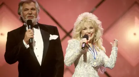Beth Gwinn/Redferns/ Getty Kenny Rogers, wearing a black and white tuxedo, and Dolly Parton, wearing a bejewelled white and cream long sleeved dress, performing on stage with a pink background.