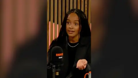 A woman in front of a BBC mic. She wears black and has long black hair.