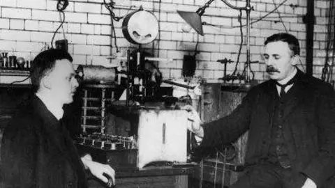 Ernest Rutherford (right) and Hans Geiger of the University of Manchester sit either side of a table of laboratory equipment in a black-and-white photograph of a physics laboratory in Manchester in 1913.