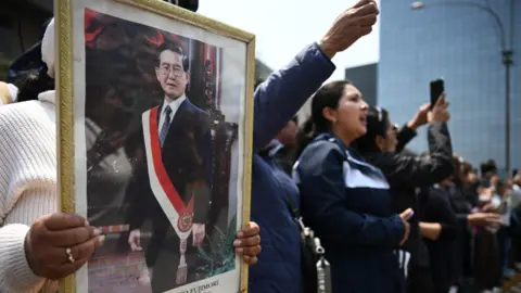 Getty Images Para pendukung mendiang Alberto Fujimori memegang bingkai foto mantan presiden yang mengenakan selempang berwarna bendera nasional Peru. Di sebelah kanan, lebih banyak orang melambaikan tangan ke udara sambil menunggu mobil jenazah Fujimori melewati kerumunan.