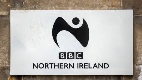 PA Media A silver plaque states BBC Northern Ireland with a black logo of a swirl and a circle. It's attached to a brick wall.