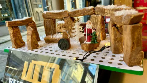 Five free-standing structures made of fudge in the shape of the Stonehenge stones, with Christmas tree and Santa ornaments in the foreground, along with a board reading Fudgehenge.