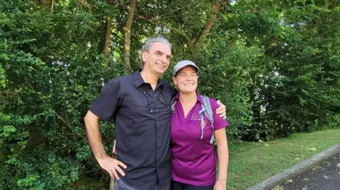 An image of Langlois and Marchand standing side by side with lush greenery behind them. He has grey hair and is wearing a black short-sleeved button up shirt with his arm around Dominique, who is wearing a cap, a backpack and a bright purple collard t-shirt. 