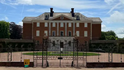 A mansion with grounds in front of it and a circular lawn. A large metal railed gate also stands in front of the house.