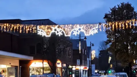 Fleet Town Council Christmas lights hanging across a busy street scene at twilight. The three of the lights are shaped like underwear, with stars and icicles also hanging down.