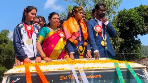 Rakshitha and her grandmother in colored clothes, along with Rahul, who is wearing a blue suit. They are standing in an open top car that is passing through a village. There is another female athlete on the left. All athletes are wearing medals. 