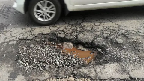 Getty Images A white car crosses a wet hole on a cracked and broken street in urban area.