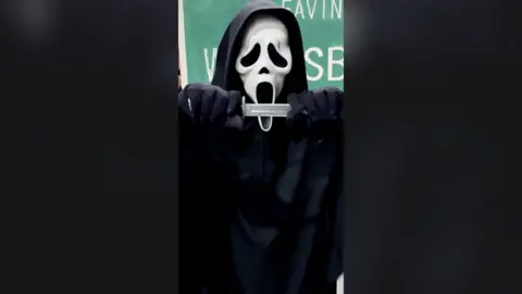 A person dressed in the costume from the Scream movies, wearing a black hooded cape and a white mask