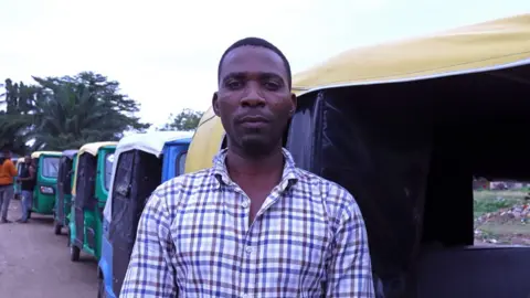 Man wearing a checked shirt stands in front of his three-wheel taxi