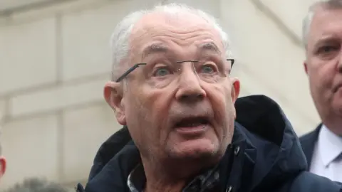 Michael McKinney - a man with short white hair wearing frameless glasses is talking outside of a white stone building.