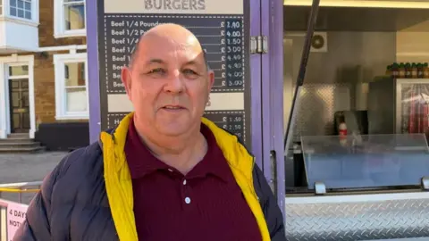 Ollie Conopo/BBC A man in a red jumper and a navy coat with yellow collars stands in front of a food truck.