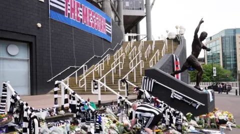 PA Media A man in a Newcastle United shirt places a bouquet on the steps St James' Park, which are covered by flowers and NUFC shirts and scarves. A flag reading The Messiah - one of the nicknames fans had for Keegan - is attached to the wall of the stadium. 