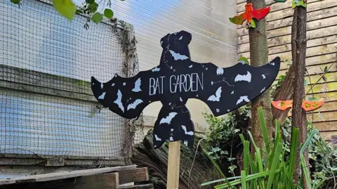 A sign is pictured within the garden. It is on a wooden stick and the sign is shaped like a bat. It reads "Bat Garden" in a large white font.