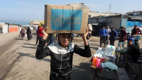 Reuters A teenage boy carries a cardboard box with the words "Unrwa" on them as dozens of people stand in the background near some toiletry supplies