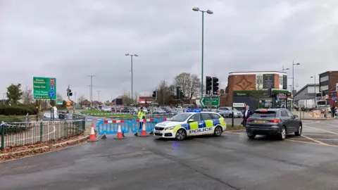 BBC A large crossroads, with road blocked off by a blue barrier and a police car. There is also a police officer standing behind the barrier and police tape blocking off the road.