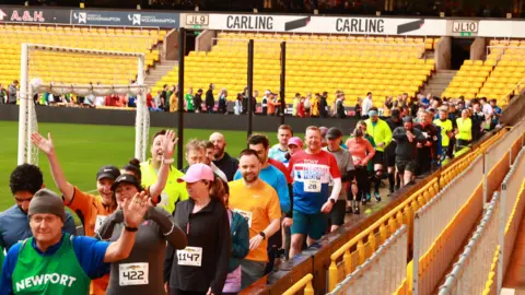 People dressed in running gear inside a football stadium that has bright yellow seating.
