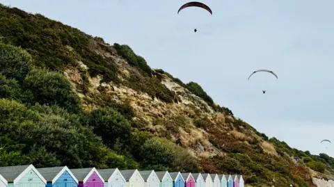agmcbride Three paragliders line up in the sky with arched canopies above beach huts and the gently sloping, vegetated cliff at Boscombe.