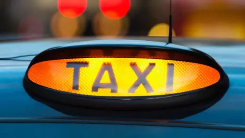 A close-up of a lit up taxi sign on the top of a black cab.