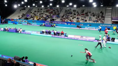 An indoor bowls contest taking place for the Commonwealth Games in Glasgow. Several players, wearing a variety of national colours, are playing at the same time, watched by a seated crowd and various officials. 