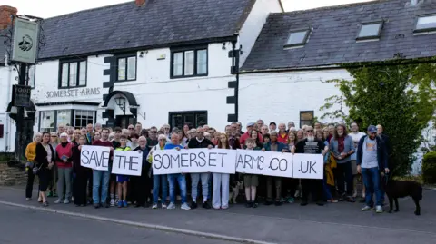 A group of people stood in front of a white building. They are holding a sign which says 'save the Somerset Arms'