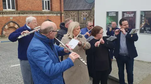 5 men and two women are standing on the street in two lines. They are all playing the flute. 