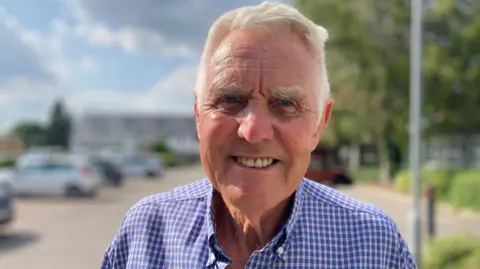 BBC/David Allard Graham Walbrin wearing a blue checked shirt standing in a car park on a bright day. Several parked cars, trees, and a cloudy sky are visible in the softly blurred background.