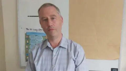 Marc McOwens Man with a blue shirt standing with his hands folded in front of him. He is an office with a wooden chair and white filing cabinet.