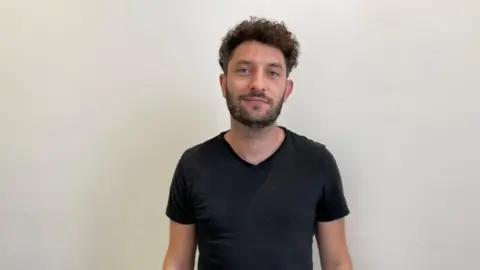 Rob Constantin/BBC Man with medium-length dark hair and beard wearing black t-shirt