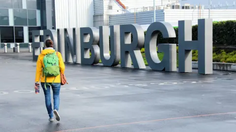 A woman wearing a yellow raincoat and green backpack walks towards the large Edinburgh lettering sign outside Edinburgh Airport.