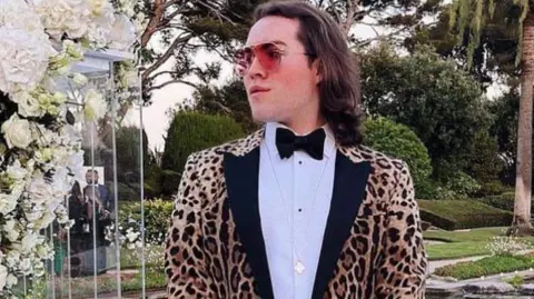 Jack Watkin is holding a handbag while wearing a leopard print jacket, white shirt and black bow tie. He is also wearing sunglasses in this social media image taken in a landscaped garden with large pond.