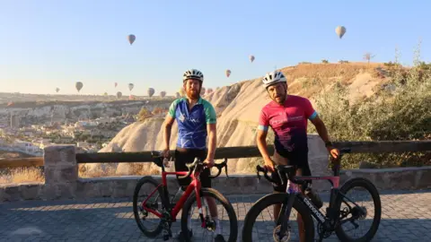 Supplied Two cyclists pose with their bikes, with hot air balloons and a Cappadocia (Turkey) landscape in the background. 