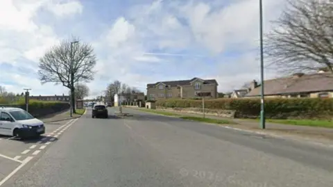 A wide paved road in a suburban or residential area during daylight.

In the foreground, the road occupies most of the image. White lane markings and a painted roadside area can be seen on the left-hand side. A small white van is partially visible at the left edge of the frame, positioned near the roadside.