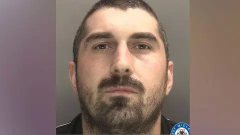 A police picture of a man looking at the camera. He has brown cropped hair, a moustache and beard. 