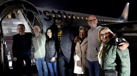 South African foreign ministry Seven people hugging and smiling in front of a plane
