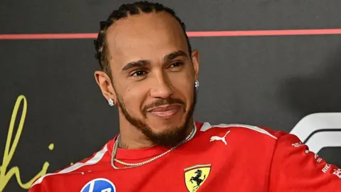Getty Images Lewis Hamilton in a red top with earrings and a necklace. He is smiling.