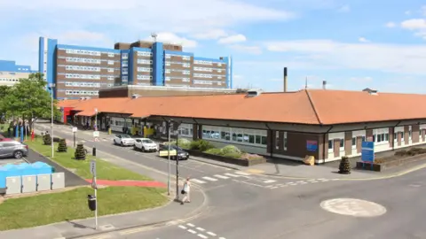 Shot of University Hospital of North Tees. It is a oblong building with a light brown slated roof. There is a block of flats in the background. There is a road around the building and cars parked beside it. 