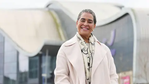 A woman with short dark grey hair, wearing a cream coat and scarf, pictured from the waist up with a glass building behind her.