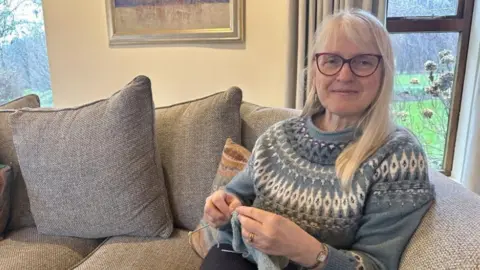 Katy Nicholls, a middle-aged woman with long fair hair, wearing spectacles and a blue Fairisle jumper. She is sitting on a sofa with grey cushions and is knitting.