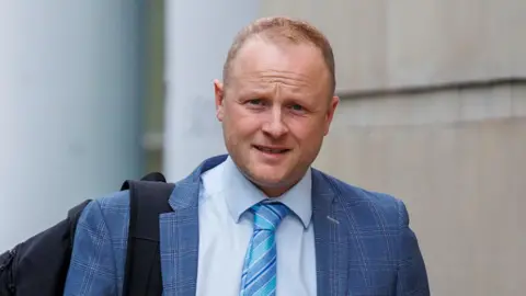 Jamie Bryson walking outside of court wearing a light blue check suit, lighter blue shirt and striped blue tie