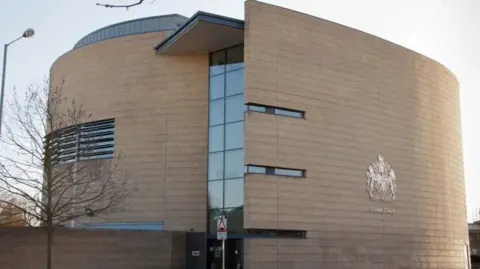 Getty Images A photo of the outside of Cambridge Crown Court. It is a circular, brick building with a strip of glass windows running down the middle of it. On the right side of the building is the Crown Court emblem. 