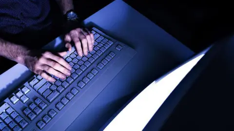 The hands of a man typing on a keyboard in front of an illuminated screen, in a darkened room