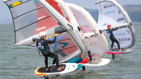 Racers on wind hydrofoil boards speed across the water from left to right. There are three of them in total.