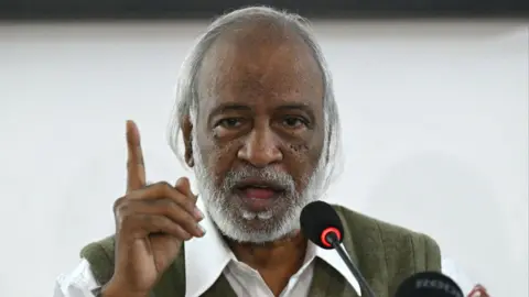 A South Asian man wearing a white shirt and green vest speaks into a microphone with a serious expression, while raising his index finger