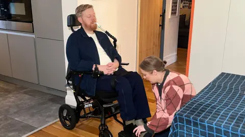 Michael Campbell is pictured in his wheelchair with his wife Naomi. He has a tube coming out of his mouth. His wife is bending down, securing his feet into straps of the wheelchair. MIchael is wearing dark clothing with a white T-shirt. His wife has on a pink and purple jumper in a diamond pattern and dark trousers. Her hair is in a bun.