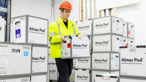 A man is wearing an orange hardhat and a yellow high-viz jacket and carrying a carboard box saying Almac Pod. There are several boxes pile don top of each other.