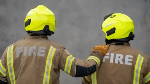 Two firefighters stand side by side in yellow and brown uniforms. One has their arm on the other's shoulder. 