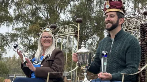 Aimee Dexter/BBC A woman and a man sitting on chairs that say Queen Conker and King Conker across the back. They are both holding silver trophies, wearing crowns and smiling. She has long grey hair and is wearing glasses, a brown coat and blue jeans. He has a dark beard and is wearing a green jumper and blue jeans.